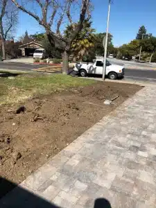 Excavated front yard area next to a paver driveway with soil piles, shovel, and parked truck on a residential street