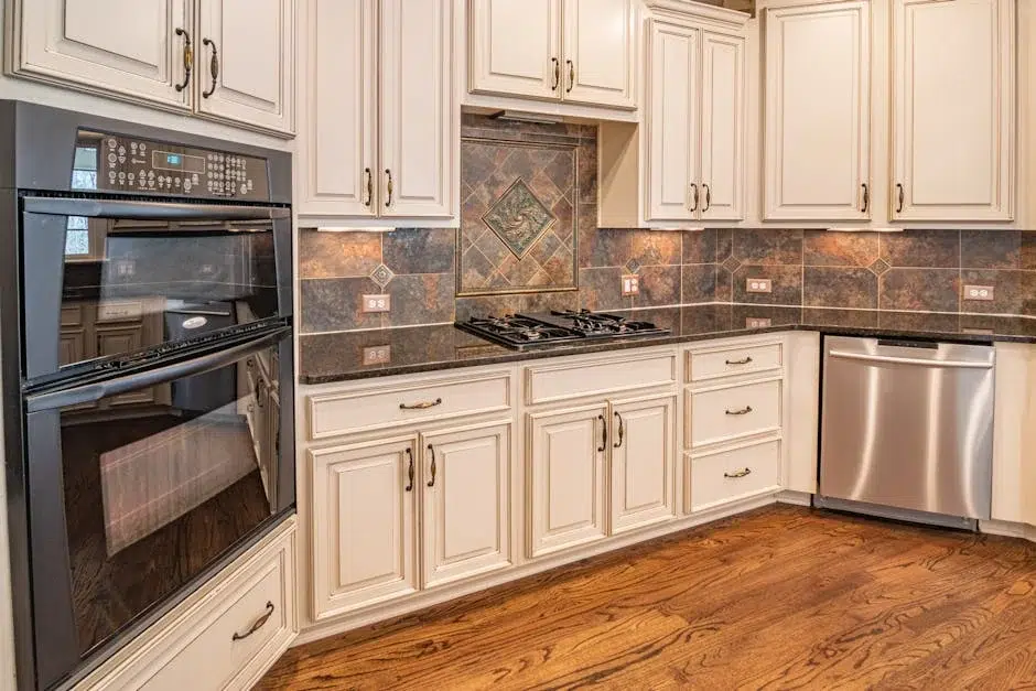 A modern kitchen featuring cream-colored cabinetry with brushed nickel hardware, dark granite countertops, and a decorative tile backsplash in earth tones. Stainless steel appliances include a gas cooktop, double wall ovens, and a dishwasher, with hardwood flooring visible throughout the space. The kitchen layout showcases an L-shaped design with ample counter space and storage.