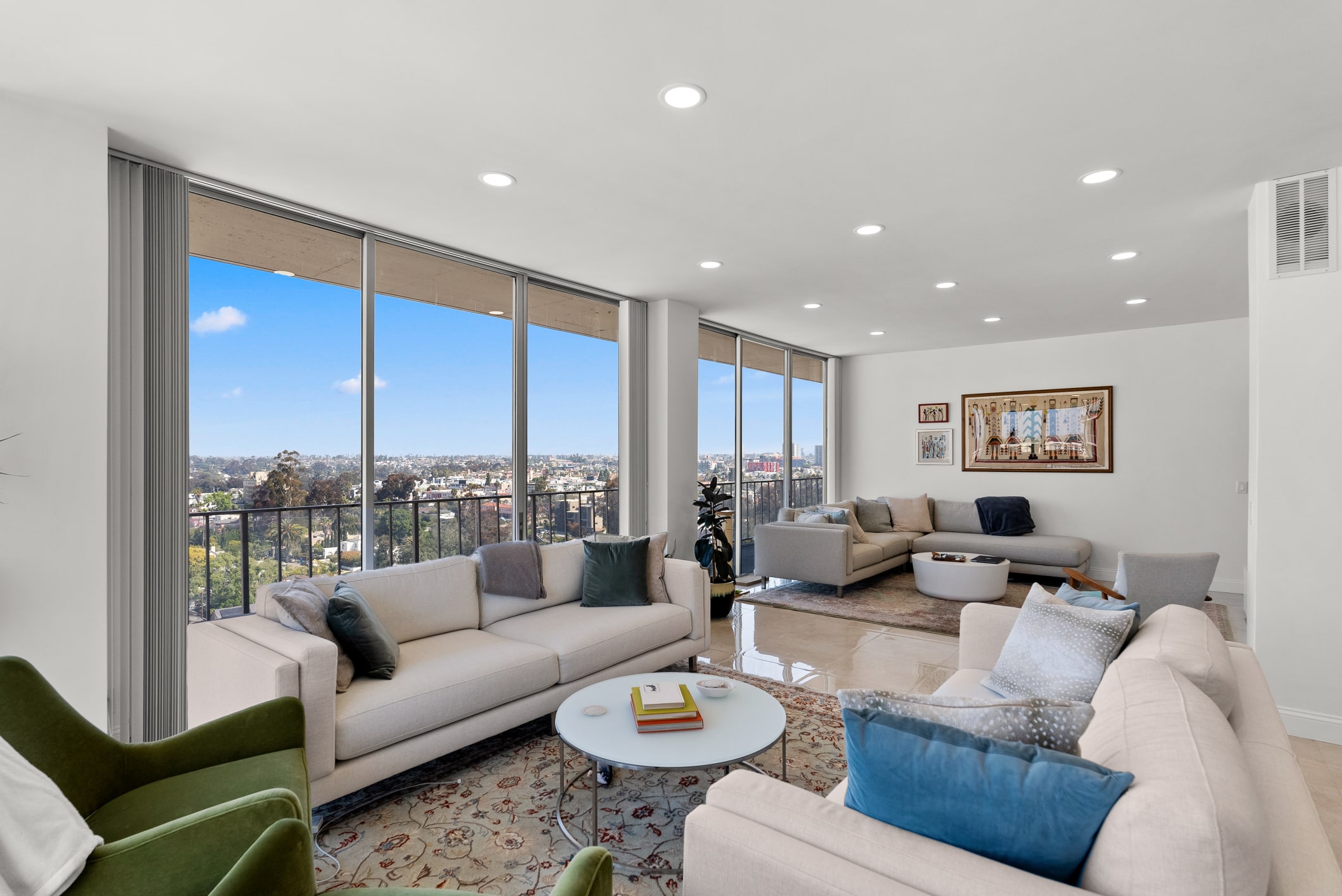 A modern high-rise living room with floor-to-ceiling windows offering panoramic city views, furnished with neutral-toned sectional sofas accented with blue and gray pillows, a white round coffee table, and green accent chairs, with recessed lighting in the ceiling and framed artwork on the walls.