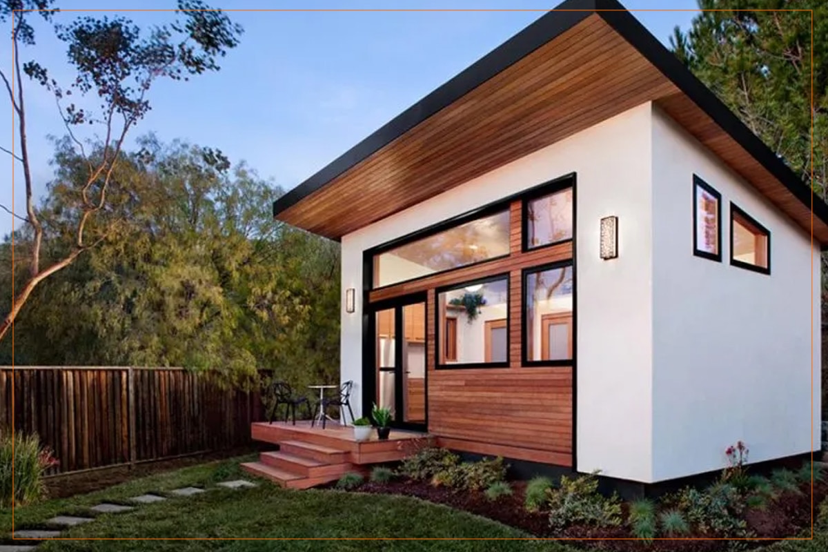A modern tiny house with white exterior walls and a dark roof features warm wood accents on the overhanging eaves and front-facing wall panels. The structure includes large black-framed windows and glass doors opening onto a wooden deck with steps, surrounded by landscaped gardens and mature trees in a natural wooded setting. The minimalist design emphasizes clean lines and sustainable architecture, exemplifying contemporary small-space living.