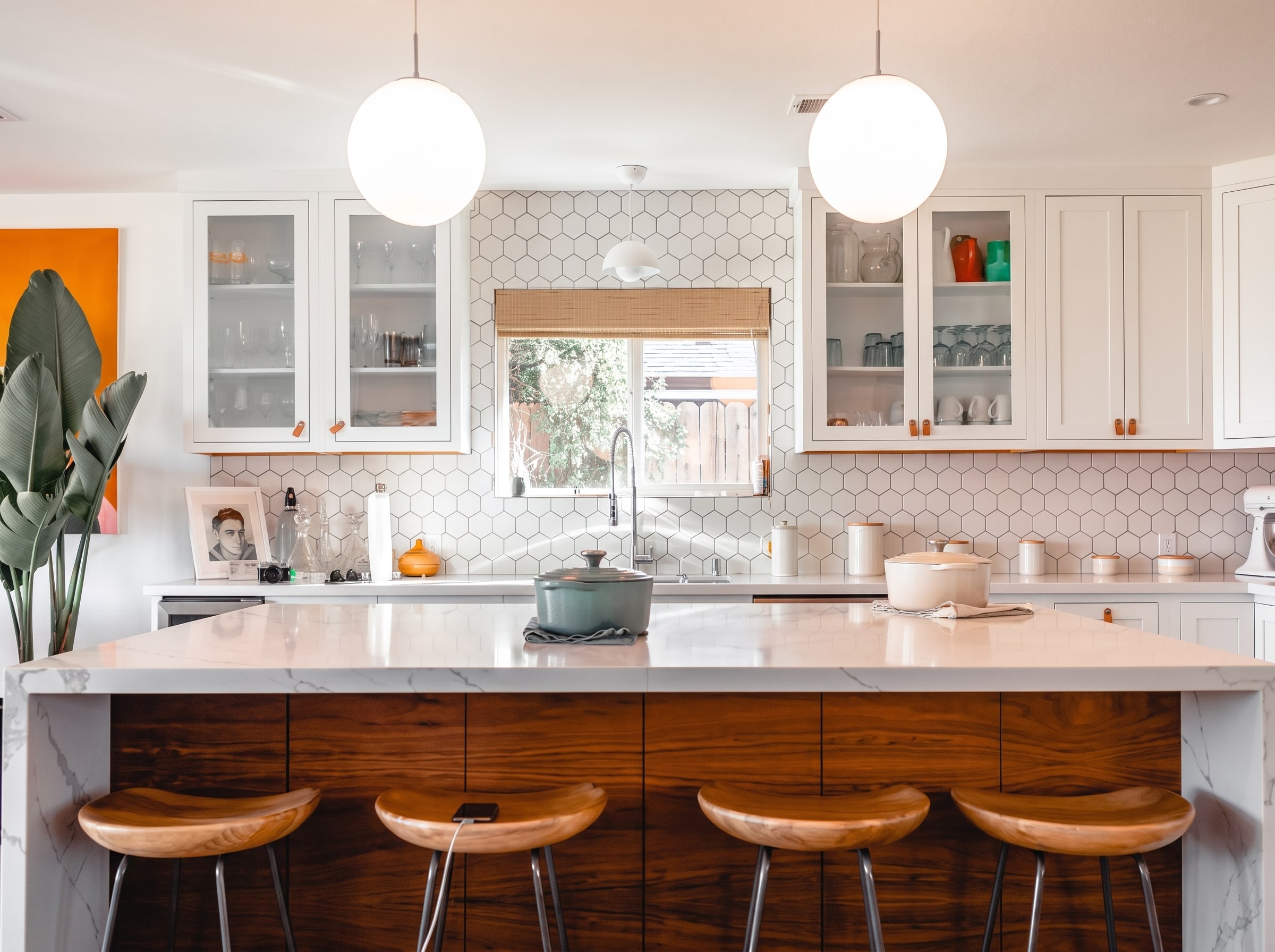 A modern kitchen features white cabinetry with glass-front doors and a hexagonal tile backsplash, complemented by a large marble-topped island with a rich wood base and four wooden mid-century style stools. Pendant lights hang above the island, while the countertops display ceramic vessels and decorative items, with a window framed by a wooden shade overlooking an outdoor space. The design combines contemporary and warm natural elements, including a potted plant on the left and orange accent details throughout the space.