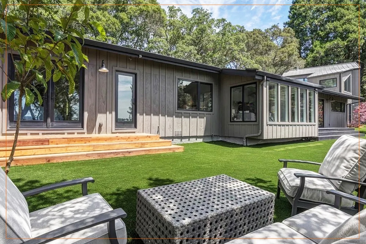 A modern residential backyard features a contemporary gray and black metal-clad home with large windows and wooden deck steps, overlooking a manicured lawn. In the foreground, comfortable outdoor seating includes upholstered chairs arranged around a woven patterned ottoman, creating a relaxed entertaining space. Mature trees frame the property in the background under a partly cloudy sky, emphasizing the home's integration with natural surroundings.