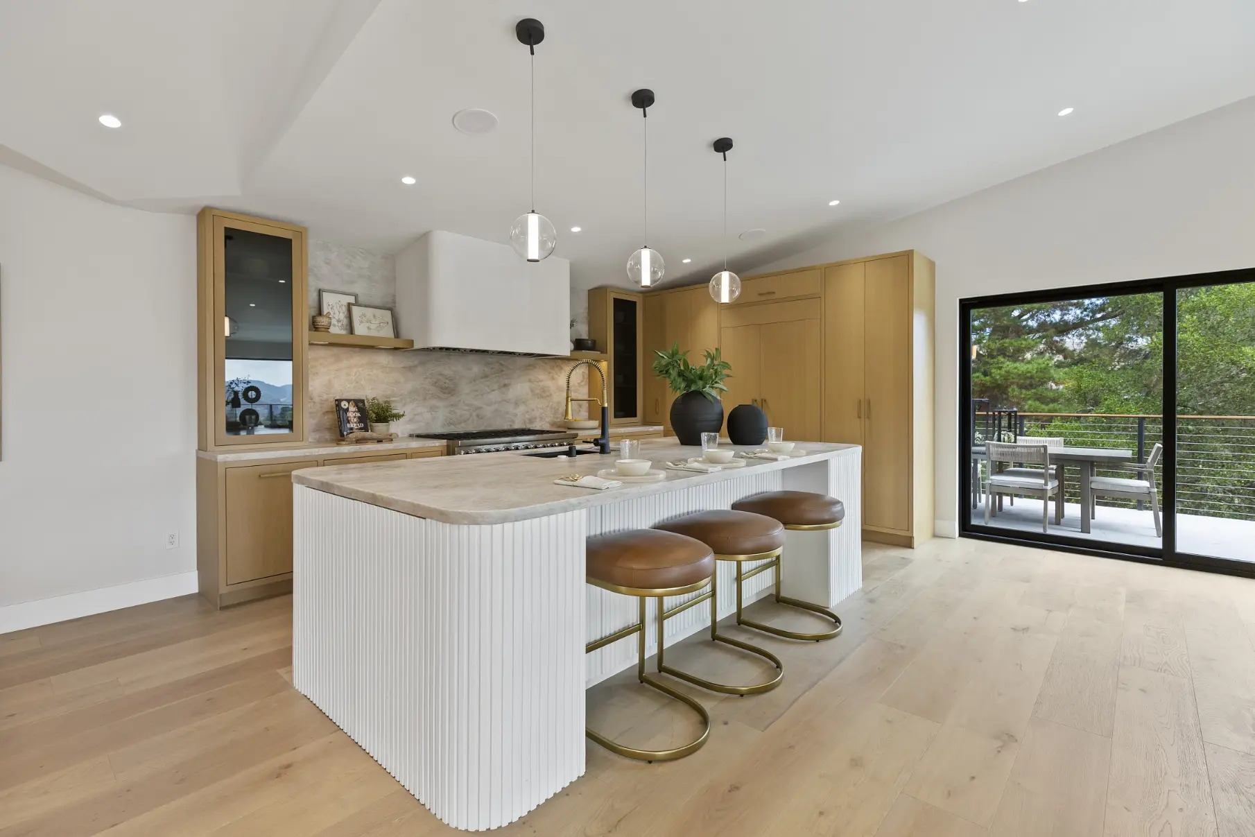 A modern kitchen featuring natural wood cabinetry in warm oak tones paired with a white ribbed island, light stone countertops, and brass and leather bar stools. The space includes pendant lighting, stainless steel appliances, and opens to an outdoor terrace visible through large black-framed glass doors, creating a bright, contemporary design with warm and neutral color palette.