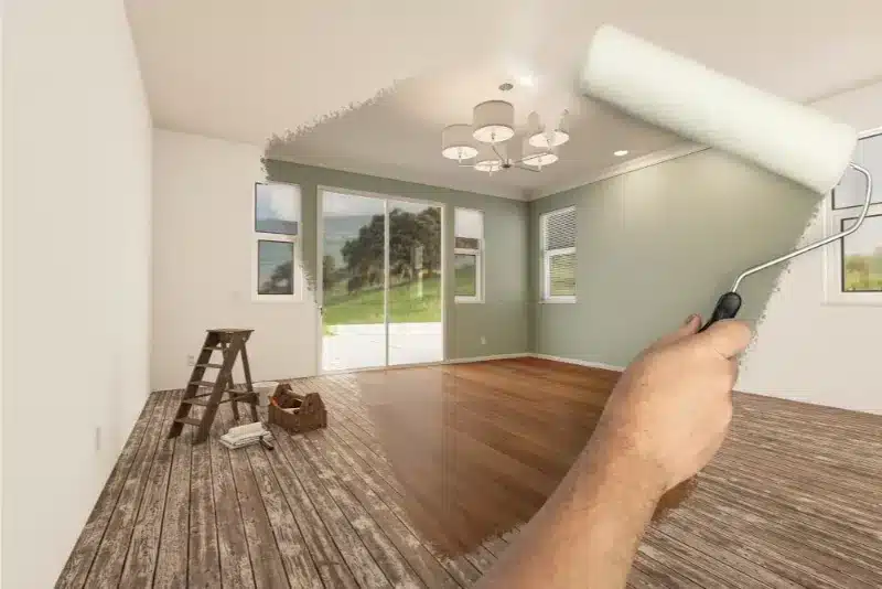 A person's hand holding a paint roller extends into a bright, spacious room undergoing renovation, with one wall partially painted sage green while others remain white. The room features hardwood flooring on one side and wood-look tile on the other, large glass sliding doors opening to an outdoor garden view, and a ladder with paint supplies positioned nearby. A modern ceiling fixture and recessed lighting are visible overhead, indicating this is a contemporary home interior renovation project.