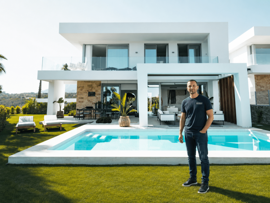 A man in a dark t-shirt and black pants stands on a manicured lawn in front of a modern white villa with a rectangular swimming pool. The contemporary two-story house features large glass sliding doors, a covered terrace area, and expansive windows overlooking scenic hillside views. The property includes lounge chairs on the grass, potted plants, and Mediterranean-style landscaping.