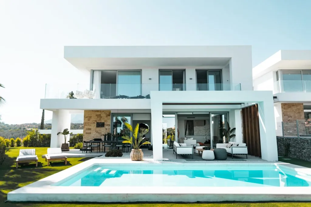 A modern white two-story residential villa with large floor-to-ceiling windows and a clean geometric design overlooks a rectangular swimming pool with turquoise water. The property features a sprawling manicured lawn, contemporary outdoor furniture, and lounge chairs positioned near the pool deck, with a stone accent wall visible on the lower level. The open-concept living areas are visible through the expansive glass doors, showcasing a minimalist interior design aesthetic in a sunny, upscale residential setting.