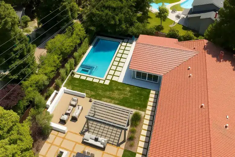 Aerial view of a residential property featuring a large swimming pool with blue water surrounded by manicured lawn and terracotta tile roofing. The estate includes a spacious patio area with lounge chairs and dining furniture, integrated seamlessly into the landscaped grounds with mature trees and greenery. The property demonstrates luxury residential architecture and design with well-maintained outdoor amenities typical of an upscale family home.