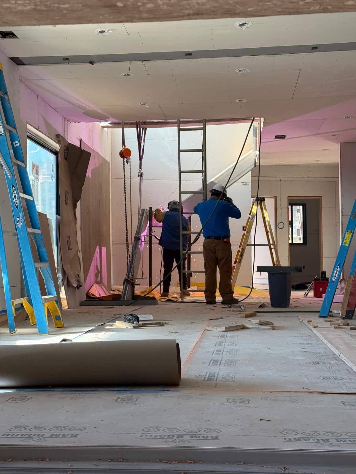 Two construction workers in hard hats and safety gear stand on scaffolding inside a commercial space undergoing renovation, with pink protective sheeting covering the walls and windows. The interior shows exposed ceiling fixtures, recessed lights, and various construction materials scattered across the floor, including rolled materials and blue ladders. The space appears to be in mid-construction with temporary containment barriers and equipment visible throughout.