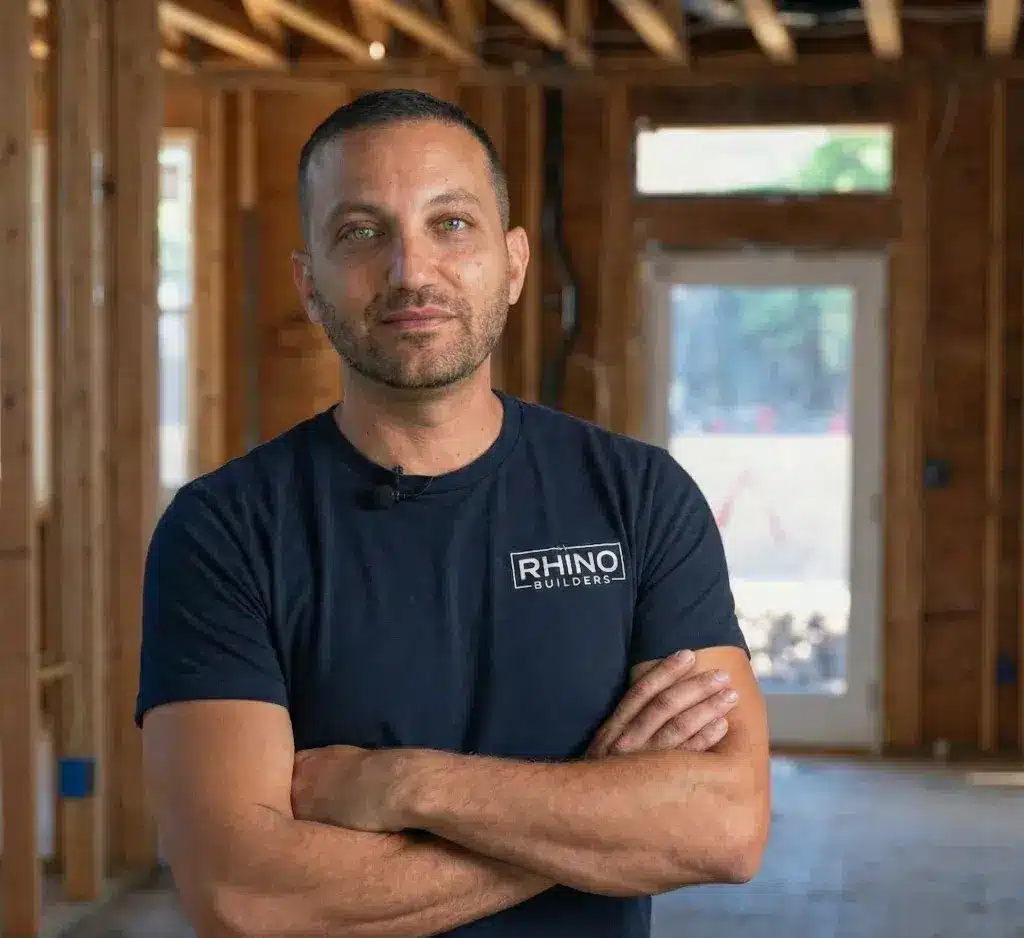A man wearing a dark navy Rhino Builders t-shirt stands confidently with his arms crossed inside an unfinished wooden building or home under construction. The interior shows exposed framing, wooden studs, and an open doorway with natural light streaming in from outside. He appears to be a construction professional or builder posing for a professional portrait.