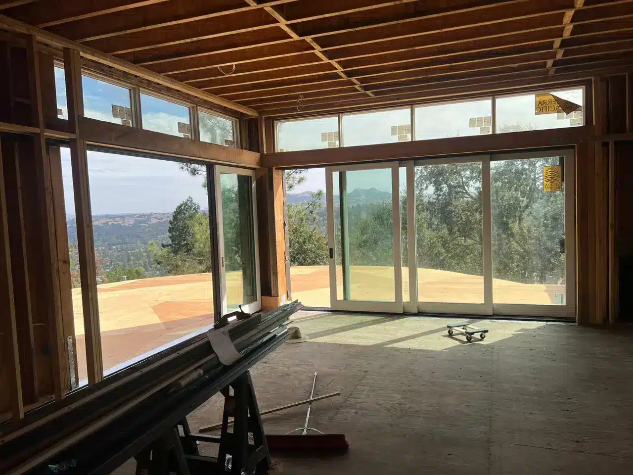 A modern residential interior under construction features extensive floor-to-ceiling glass doors and windows framing panoramic views of rolling hills, forested valleys, and distant mountains. The exposed wood beam ceiling and concrete subfloor indicate the space is in mid-renovation, with construction equipment visible on the floor. The expansive glazing opens onto a terracotta-tiled deck, creating a seamless indoor-outdoor living space designed to maximize natural light and scenic vistas.