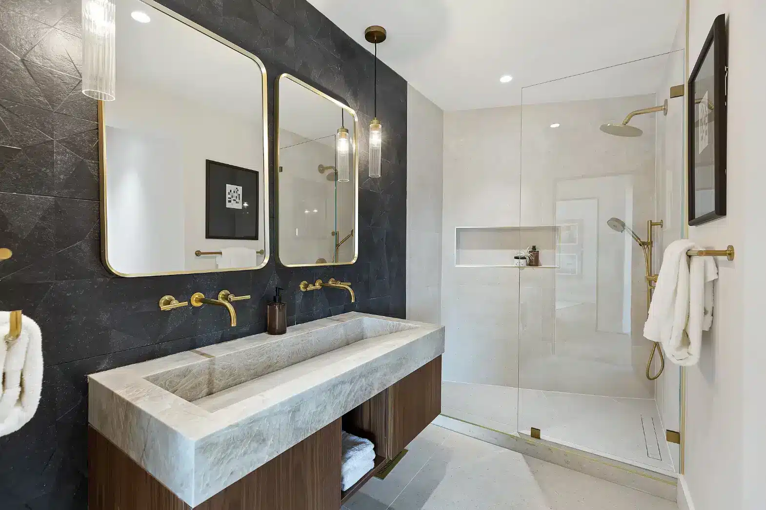A modern luxury bathroom features a dark slate accent wall with two large gold-framed mirrors and brass wall-mounted faucets above a white marble trough sink. To the right, a spacious walk-in shower with glass enclosure and brass fixtures is complemented by white subway tile walls and a rainfall showerhead. The space is accented throughout with brass hardware, pendant lighting, and white towels, creating an elegant contrast between the dark and light design elements.