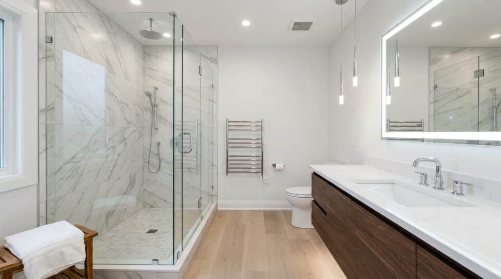 A modern, bright bathroom features a spacious walk-in shower with marble tile walls and a glass enclosure, positioned on the left side of the room. The right side displays a dark wood vanity with a white countertop, an illuminated mirror with integrated lighting, and chrome fixtures, while warm wood-look flooring runs throughout. Additional amenities include a heated towel rack, pendant lighting, recessed ceiling lights, and a toilet, creating a luxurious spa-like atmosphere.
