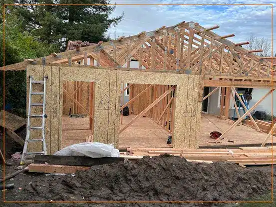 A residential house under construction shows the wooden framing and structural skeleton of the building in progress. The exposed frame reveals the interior wall studs, roof trusses, and window openings, with insulation material visible in some sections. The construction site includes building materials and equipment scattered around the foundation, indicating active development of this residential project.