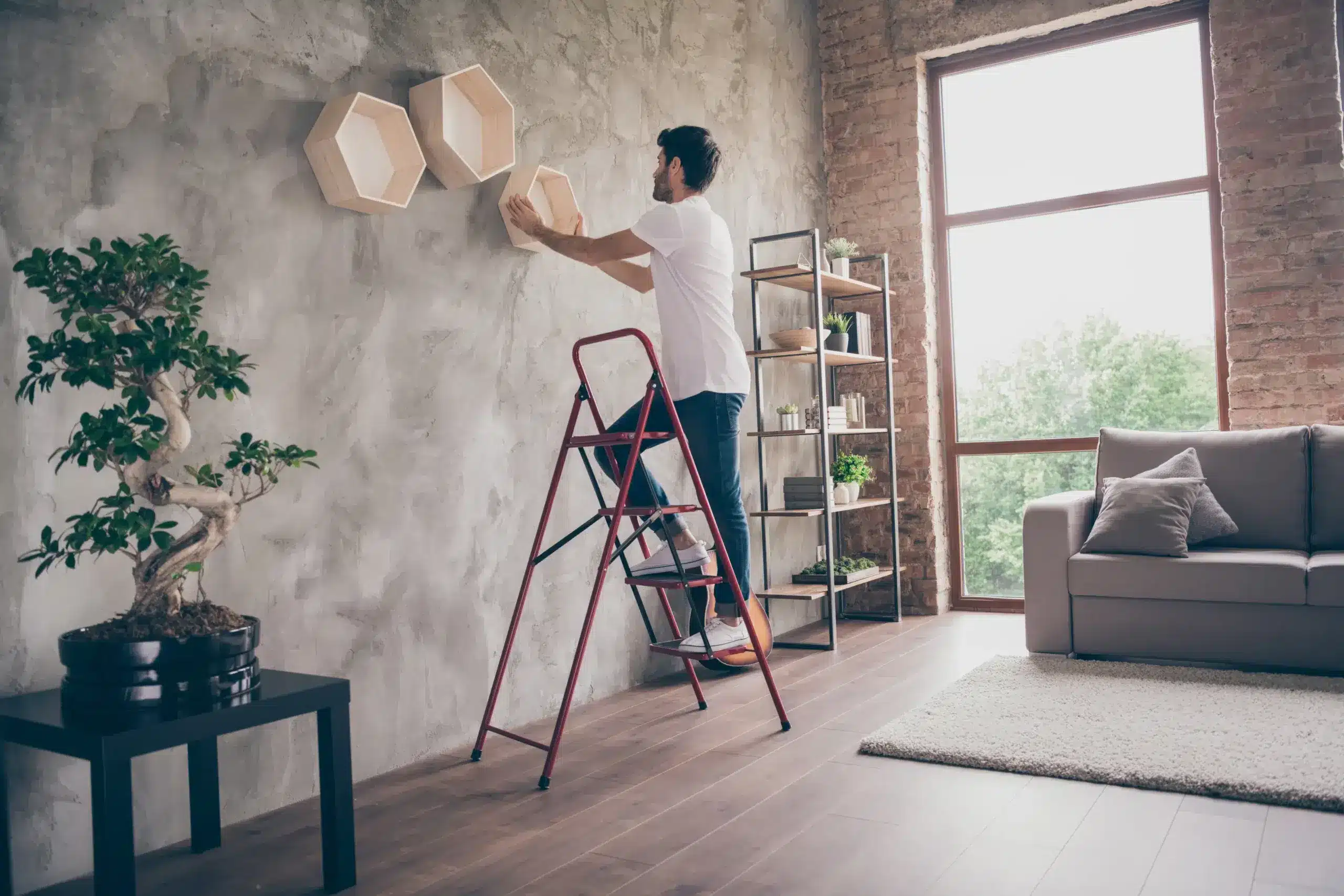 A man stands on a red ladder hanging geometric wooden wall art on a concrete accent wall in a modern loft apartment. The spacious room features exposed brick, large windows with natural light, a gray sofa with pillows, potted plants including a bonsai tree, and industrial shelving. The contemporary interior design emphasizes minimalist décor and natural elements.