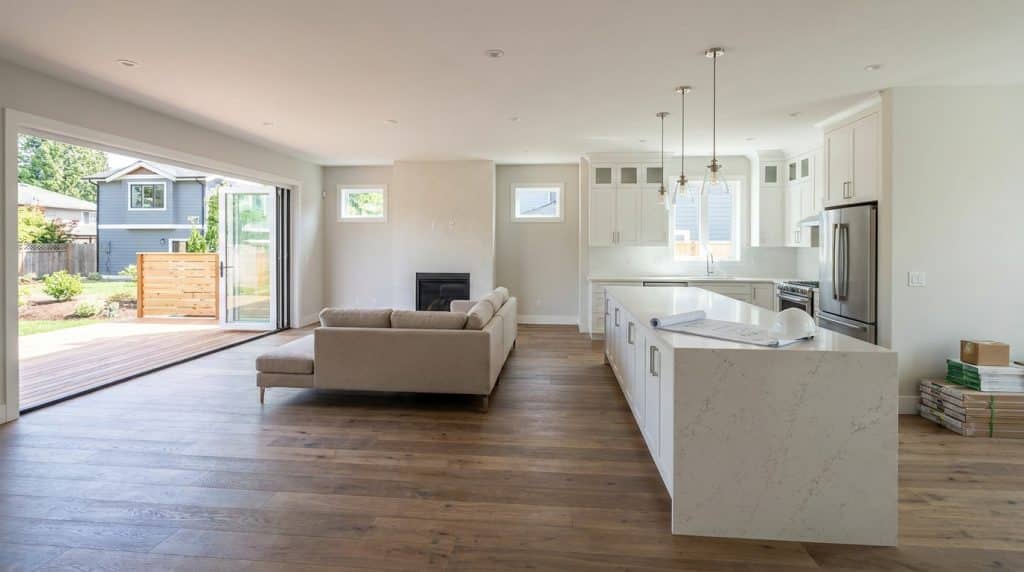 A modern, open-concept home interior featuring a spacious living area with a beige sectional sofa, a kitchen with white cabinetry and quartz countertops, and hardwood flooring throughout. Large glass sliding doors open to a backyard patio and deck, while pendant lights hang above the kitchen island and a fireplace sits centrally between the living and kitchen spaces. The bright, airy design emphasizes clean lines and neutral tones with stainless steel appliances.