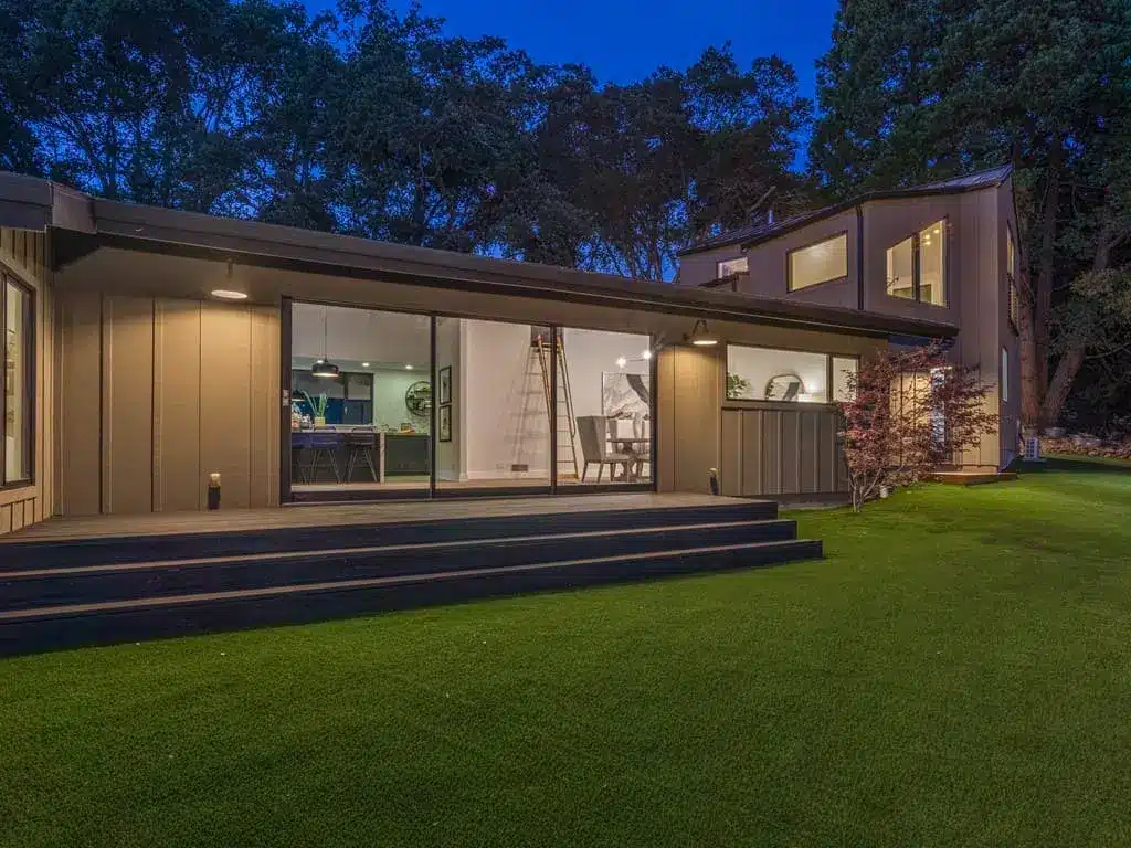 A modern residential dwelling photographed at twilight features a low-profile design with extensive glazing and warm interior lighting. The single-story home showcases clean lines, dark exterior cladding, and a raised deck with dark steps facing a manicured lawn. Mature eucalyptus trees frame the property against a deepening blue evening sky, creating a contemporary architectural contrast with the natural landscape.