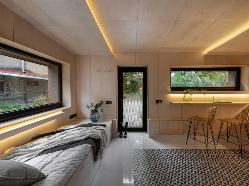 A modern interior space combining a bedroom and kitchenette, featuring warm wood paneling walls, ambient LED lighting along the ceiling and window sills, and a bed with gray bedding positioned beneath a large window overlooking greenery. The open-plan layout includes a kitchen counter with two rattan bar stools, a patterned geometric floor, and multiple windows and a door providing natural light and access to an outdoor garden area.
