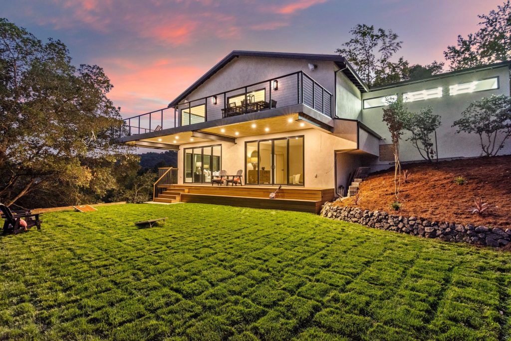 A modern two-story residential home with clean architectural lines sits on a sloping hillside during dusk, featuring illuminated large windows, a wooden deck with metal railings, and a cantilevered upper level. The property showcases a manicured green lawn in the foreground, surrounding landscaping with native plants and stone features, and is nestled among mature trees with hills visible in the background. Warm interior and exterior lighting highlights the contemporary design as the sky displays soft pink and purple sunset hues.