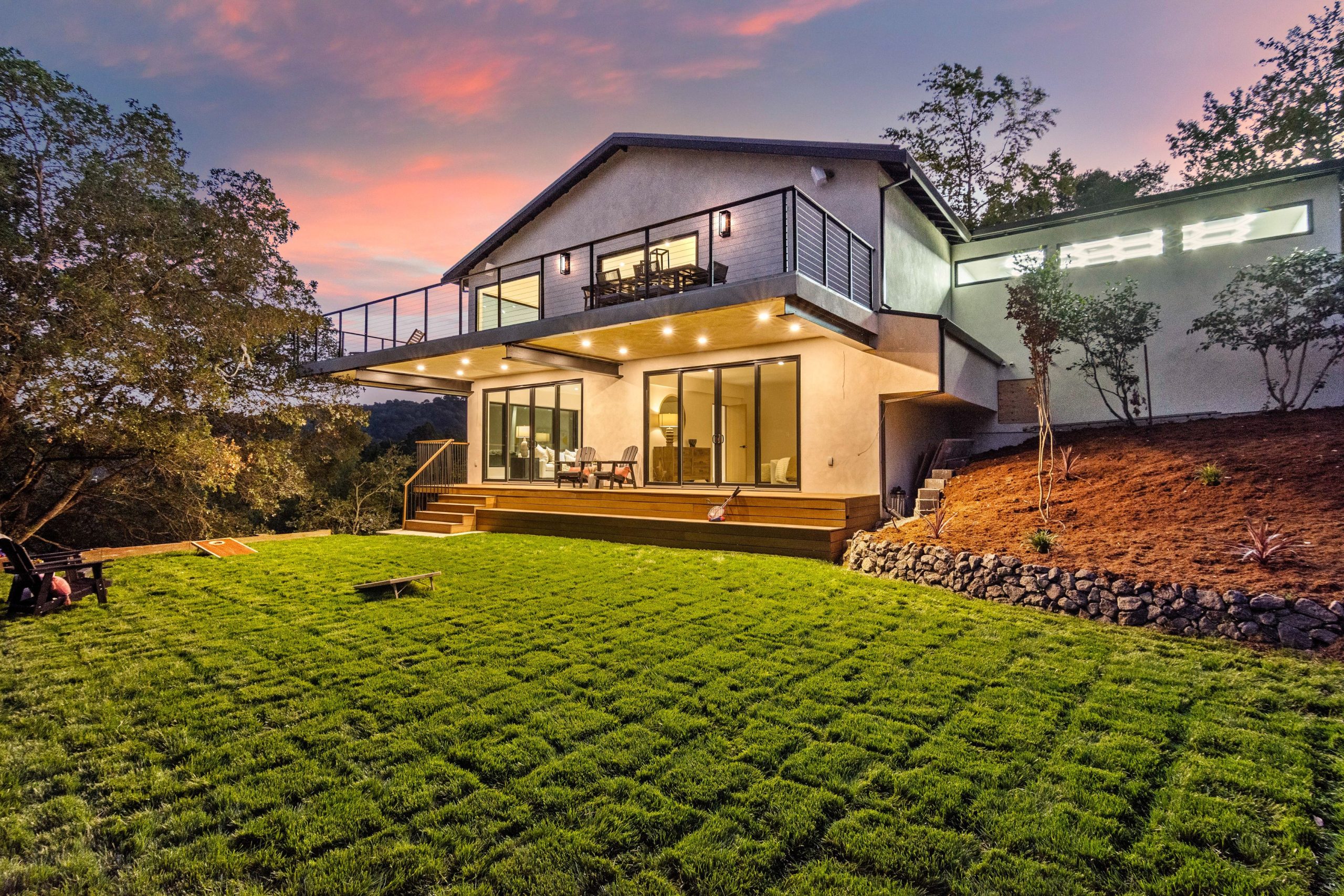 A modern two-story residential home with clean architectural lines sits on a sloping hillside during dusk, featuring illuminated large windows, a wooden deck with metal railings, and a cantilevered upper level. The property showcases a manicured green lawn in the foreground, surrounding landscaping with native plants and stone features, and is nestled among mature trees with hills visible in the background. Warm interior and exterior lighting highlights the contemporary design as the sky displays soft pink and purple sunset hues.