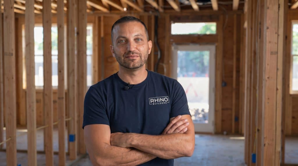 A man wearing a dark navy t-shirt with "RHINO BUILDERS" branding stands with his arms crossed inside an unfinished wooden-framed house under construction. He is positioned in a hallway or interior space with exposed wooden studs, framing, and windows visible in the background, appearing confident and professional in the construction setting.