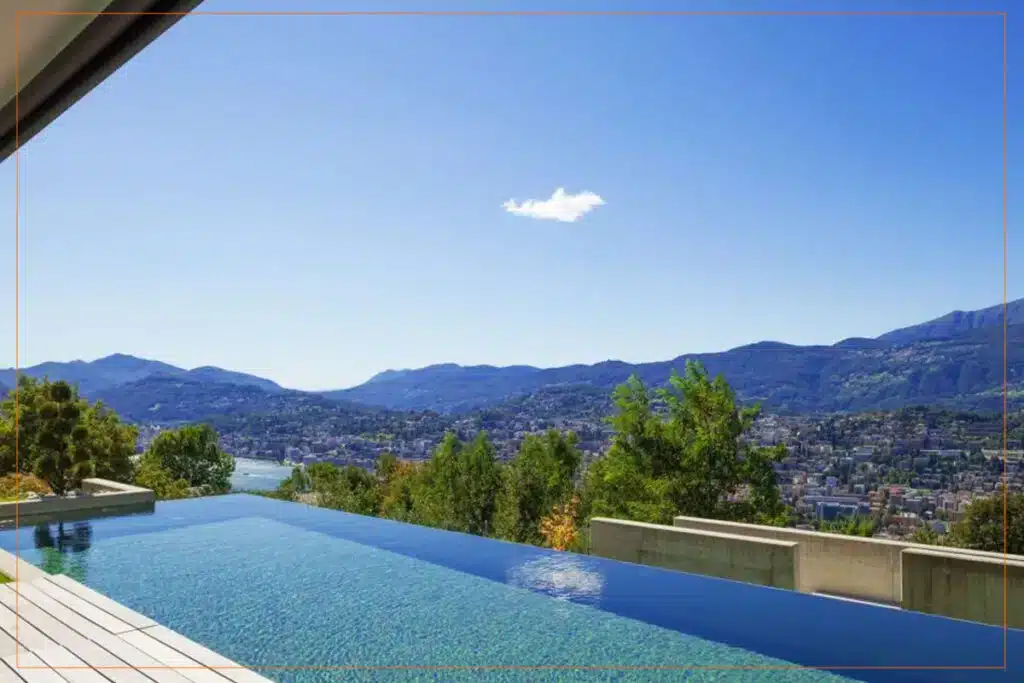A luxurious infinity pool on a modern hillside terrace overlooks a picturesque lake and mountain valley dotted with residential buildings. The crystal-clear turquoise water of the pool blends seamlessly with the distant blue waters and sky, while lush green trees frame the contemporary architecture. Bright sunshine illuminates the serene landscape under a mostly clear blue sky with wispy clouds.