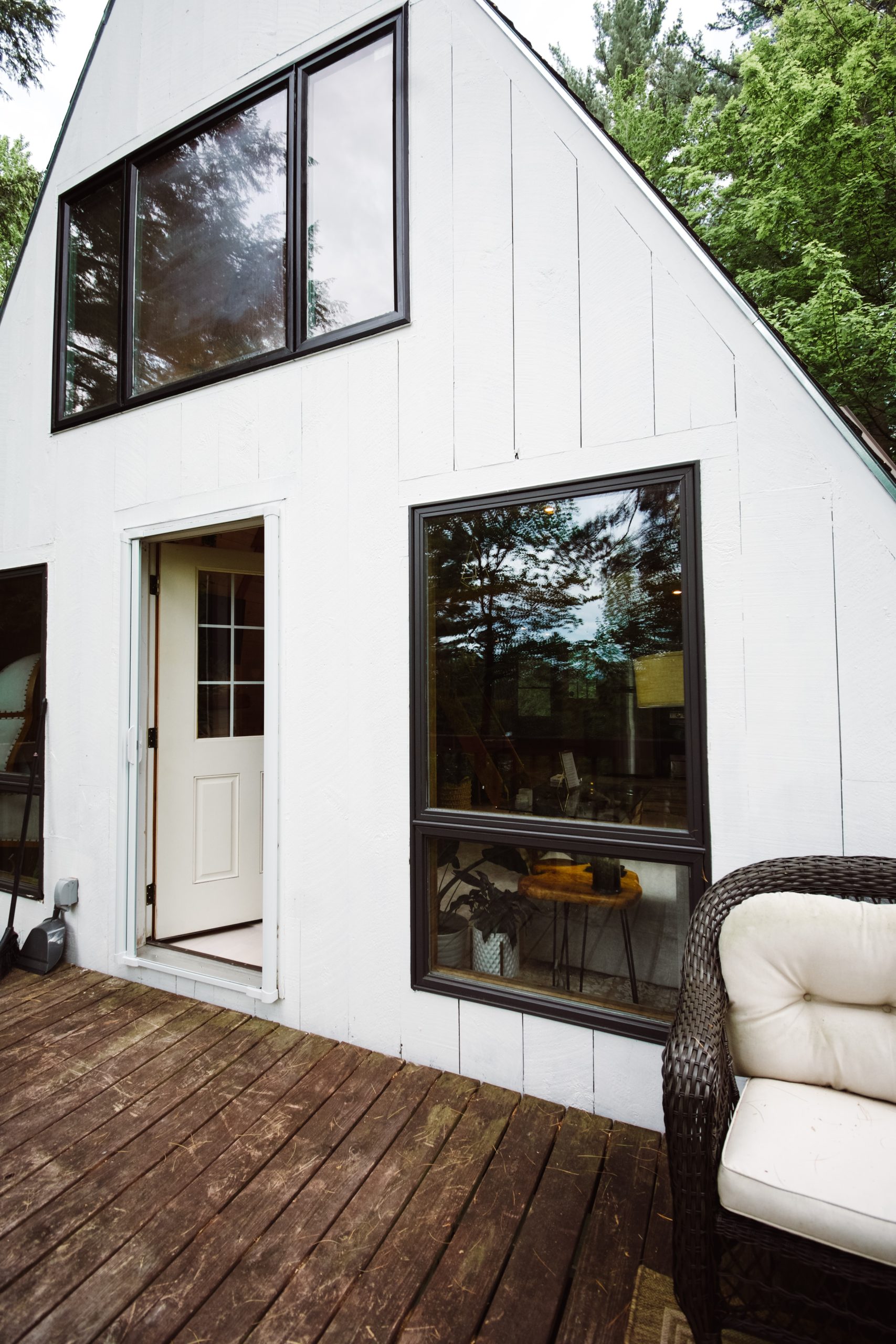 A modern white residential building with black-framed windows and a wooden deck features a white entry door and large glass panels. The structure is surrounded by lush green trees and vegetation, creating a natural woodland setting. Outdoor furniture with cream-colored cushions is visible in the foreground on the deck.