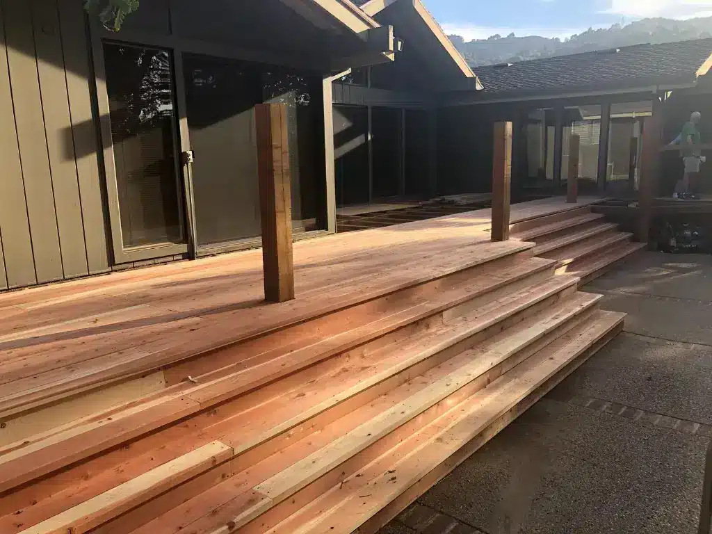 A spacious wooden deck under construction or renovation, featuring freshly laid timber planks in varying shades of natural wood tones. The deck is attached to a modern residential home with large glass sliding doors and black-framed windows, supported by wooden posts. Mountains are visible in the background under a partly cloudy sky, with the afternoon sun casting long shadows across the deck surface.