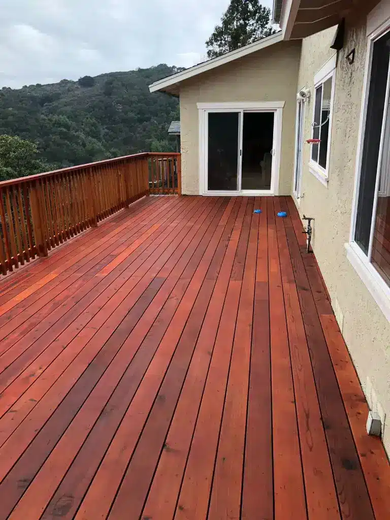 A spacious wooden deck with a rich reddish-brown stain extends from a cream-colored house, featuring white-framed glass doors and matching windows. The deck is enclosed by a wooden railing and overlooks a lush, forested hillside with dense green vegetation. Two blue objects are visible on the deck surface, and the sky above shows overcast conditions.