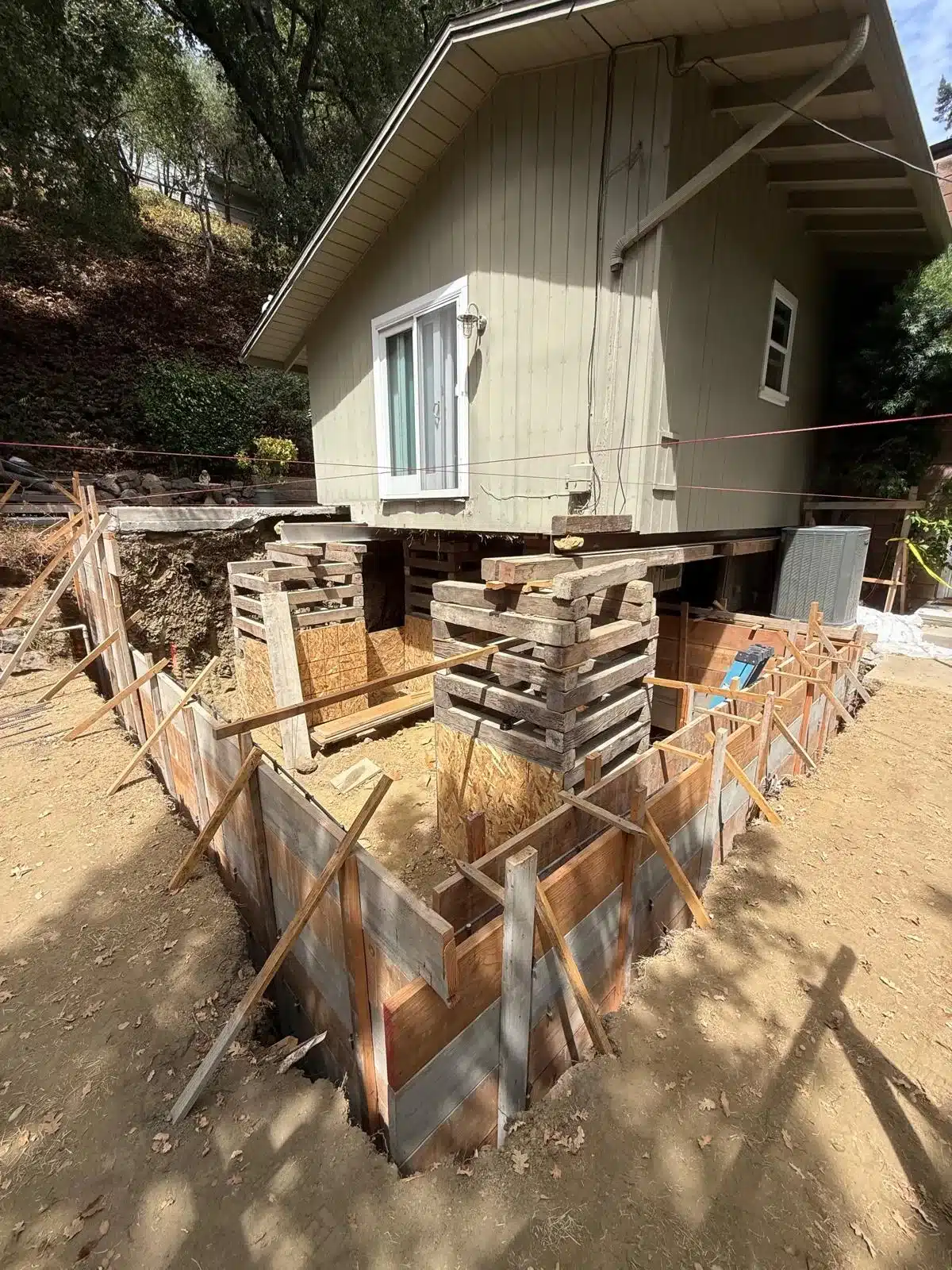 House supported on stacked timber cribbing above excavated ground, with wood forms and braces around the opening.