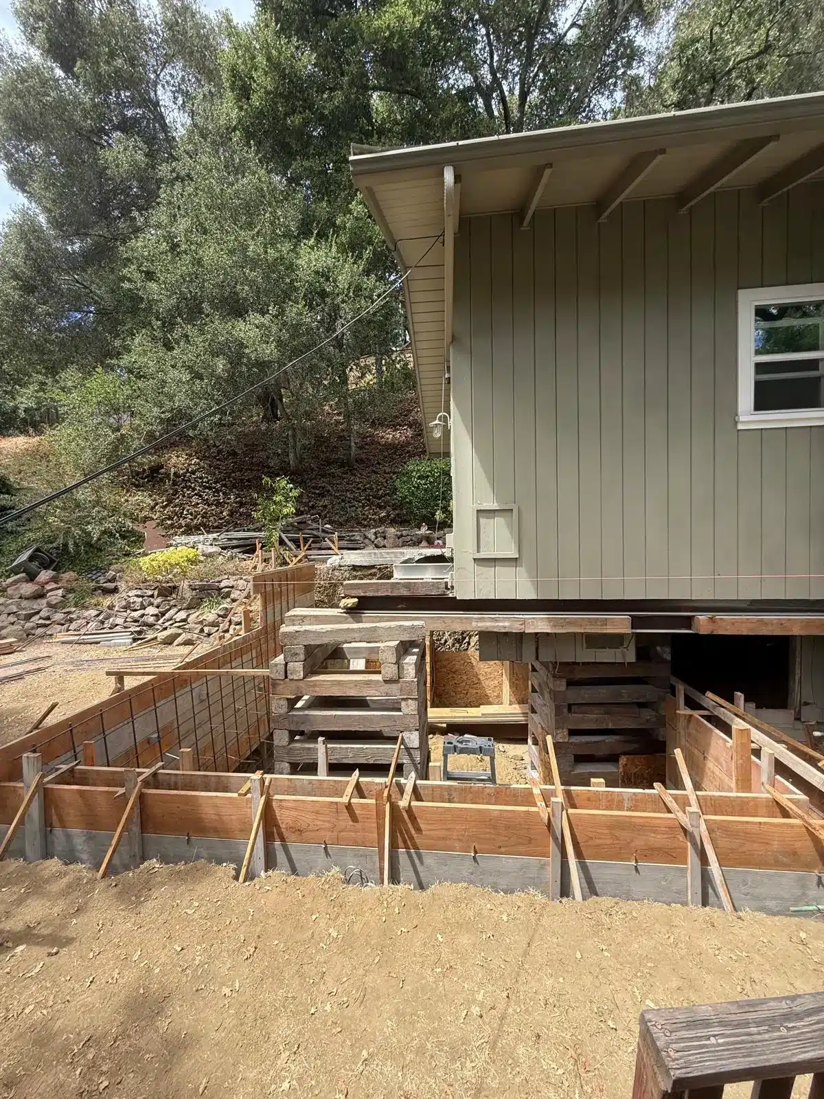 Side view of a house on timber cribbing above an excavation, with wood concrete forms and reinforcing steel visible.