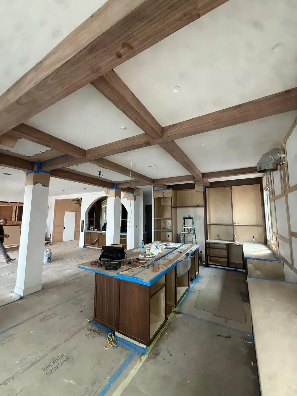 Grid of wood ceiling beams above island cabinetry, with protective paper, blue tape, tools, and building materials.