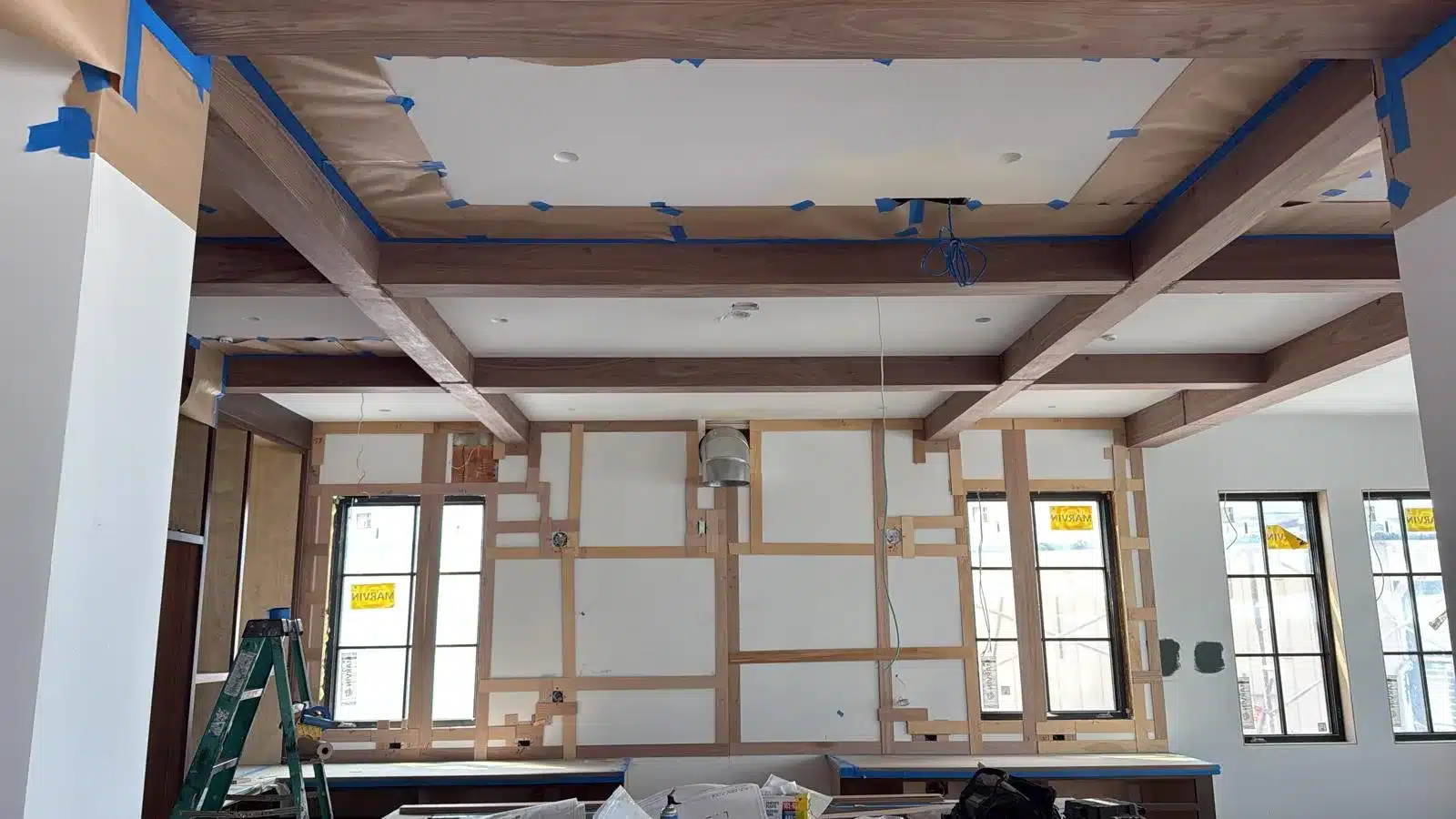 Wood ceiling beams and unfinished wall trim around black-framed windows, with masking paper, blue tape, and a ladder.