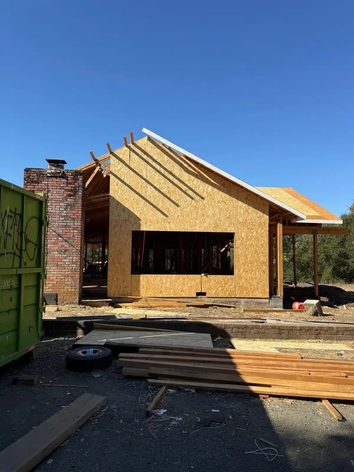 House under construction with exterior sheathing, exposed roof framing and unfilled window openings beside a brick chimney.