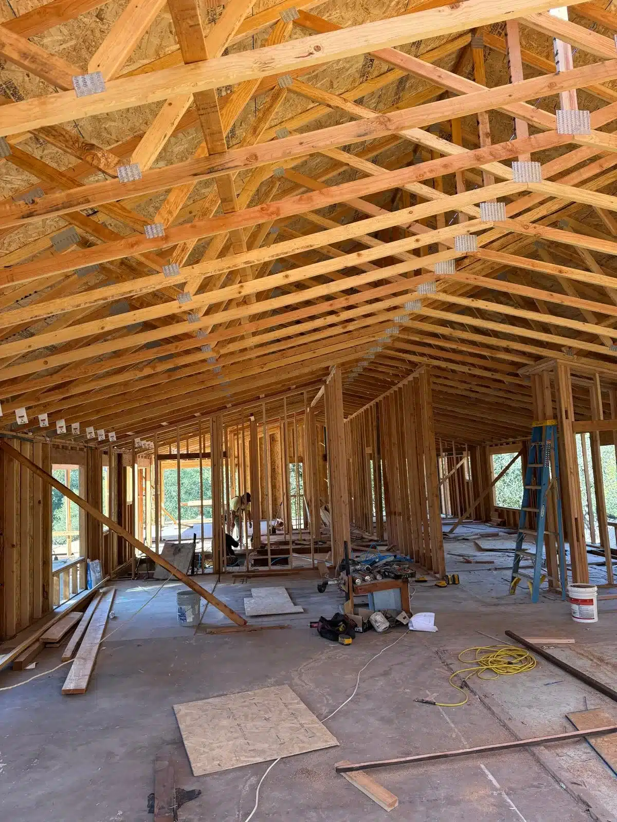 Home interior during framing with exposed roof trusses, metal connectors, wall framing and construction tools on the floor.