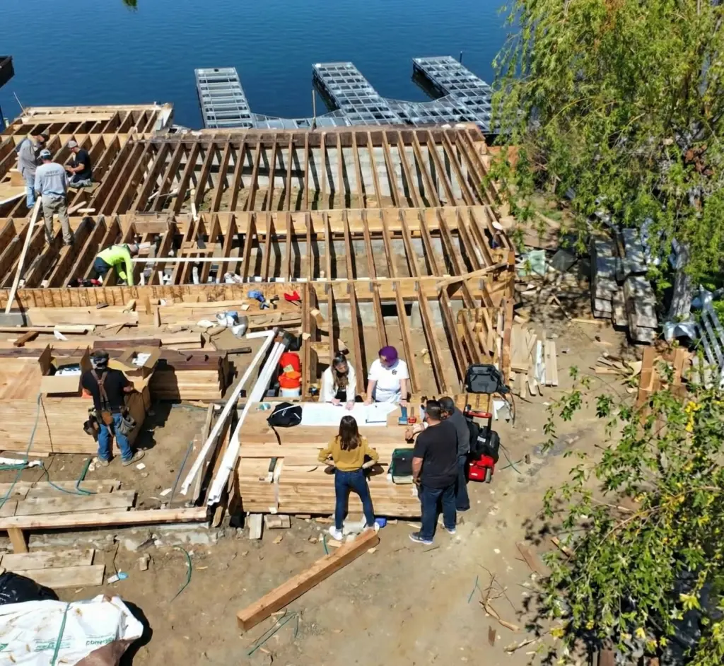 Construction crew reviewing plans on a wooden deck frame under construction beside a lake with docks in the background