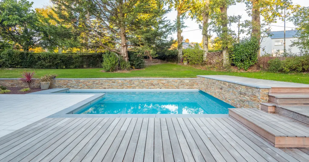 Modern backyard pool with wood deck, stone retaining walls, and landscaped lawn surrounded by mature trees