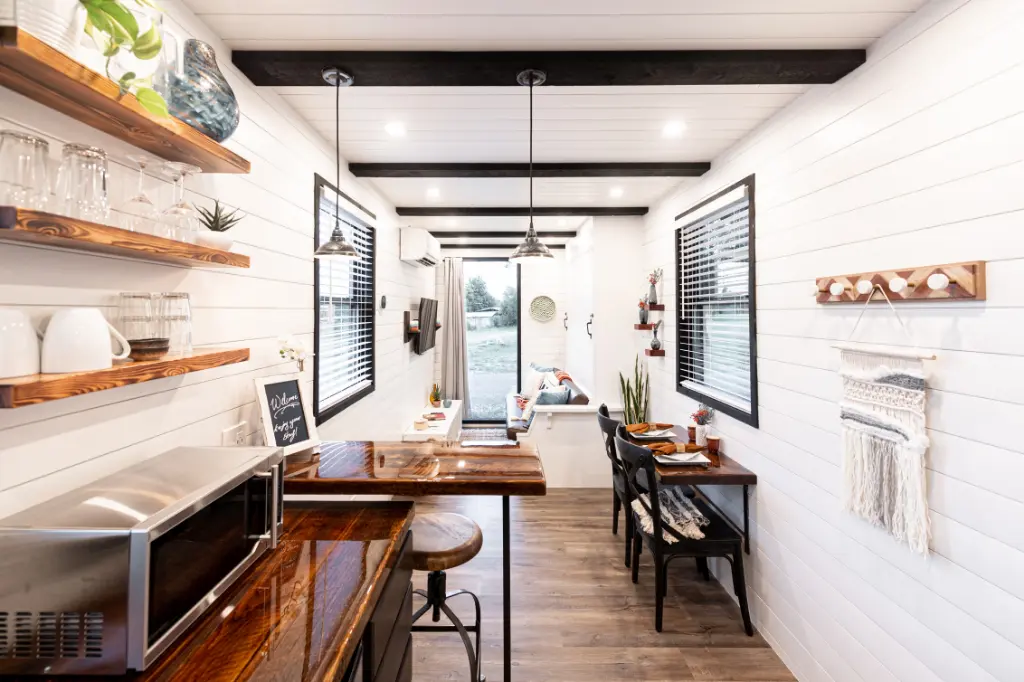 Narrow modern kitchen and dining space with white shiplap walls, open shelving, wood counters, and black beams