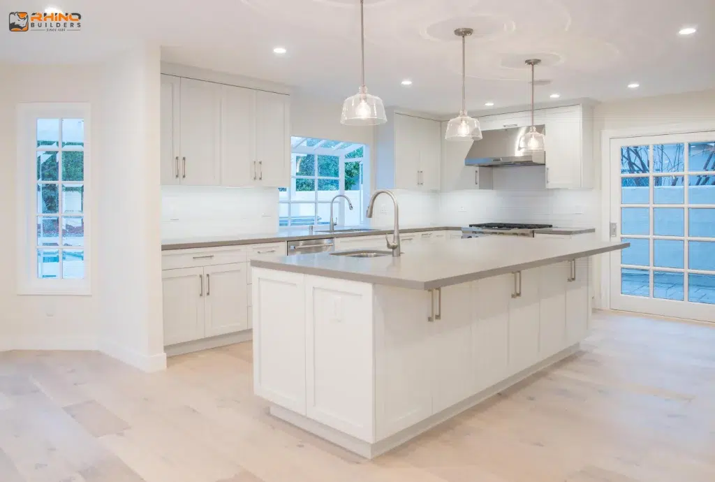 All-white kitchen with shaker cabinets, large island, pendant lights, and stainless appliances near glass patio doors.
